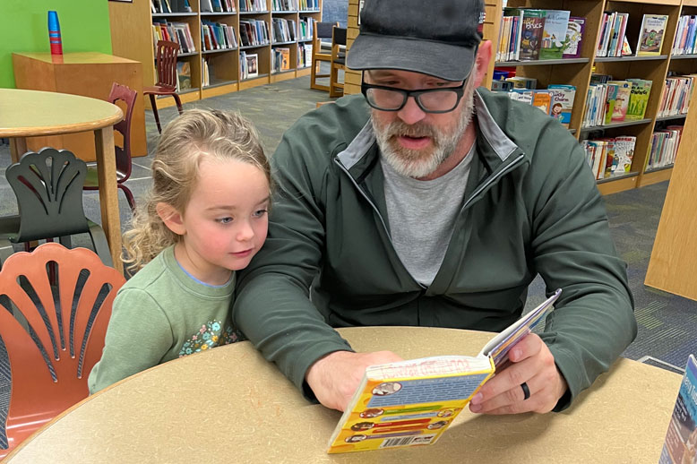 Father and child sit at a library table reading a book