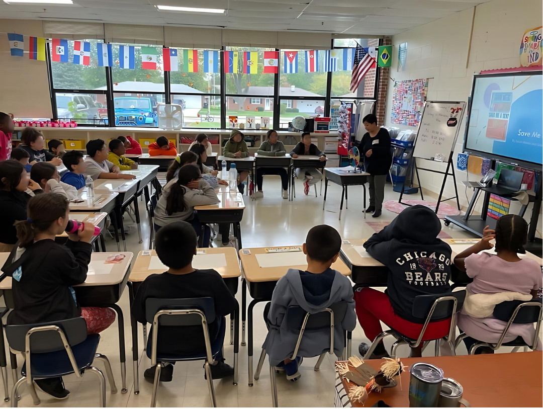 Library staff member stands at the front of a classroom with elementary aged students at their desks.