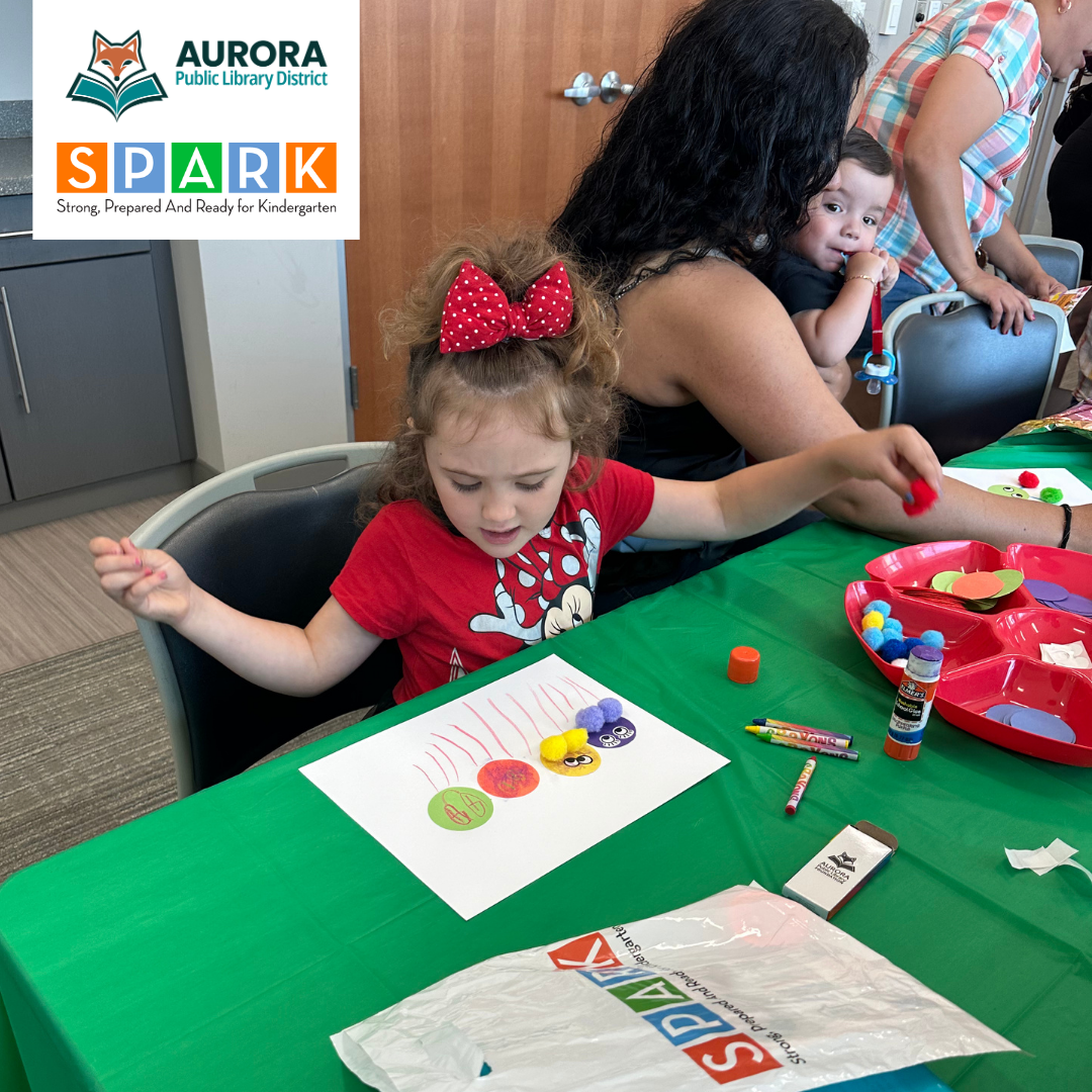 A young child sits at a table an works on a craft activitis with colorful paper, pom poms, and glue during a library program with families.